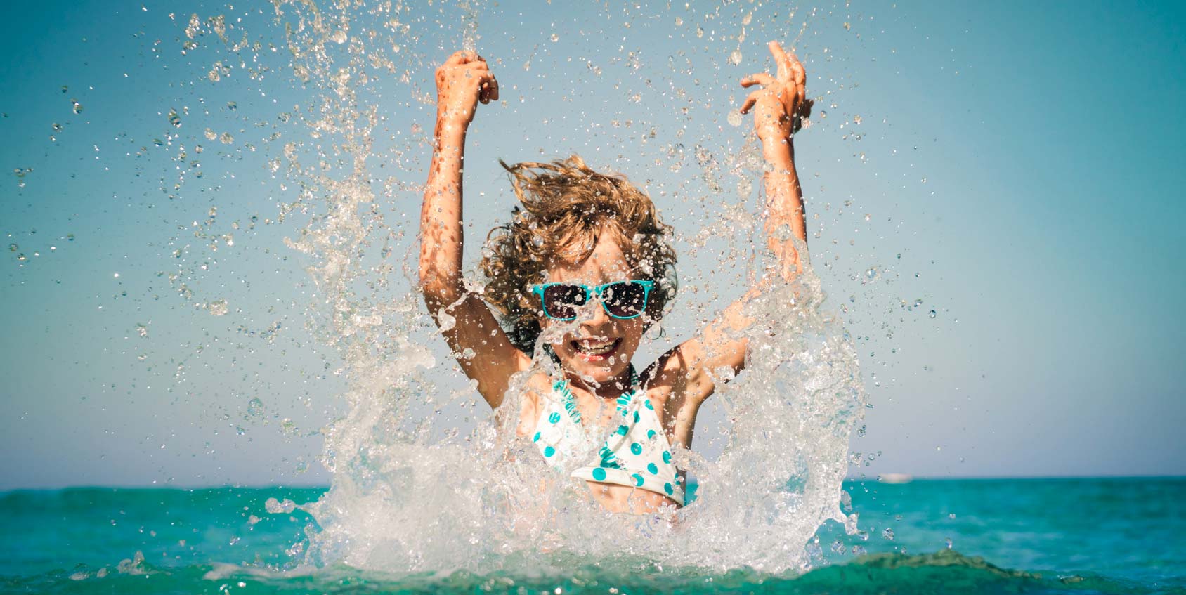 Jeune fille dans l'eau au bord de mer les bras en l'air avec des lunettes de soleil, éclabousse l'eau autour d'elle